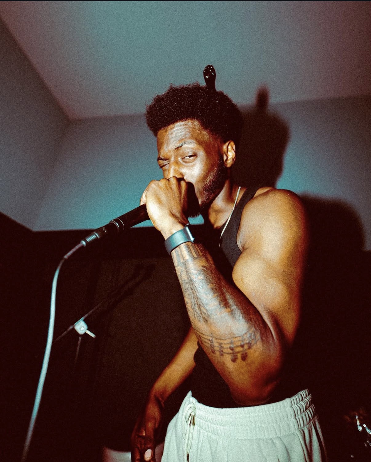 Male performer singing into a microphone, with a tattooed arm, in a dimly lit room.