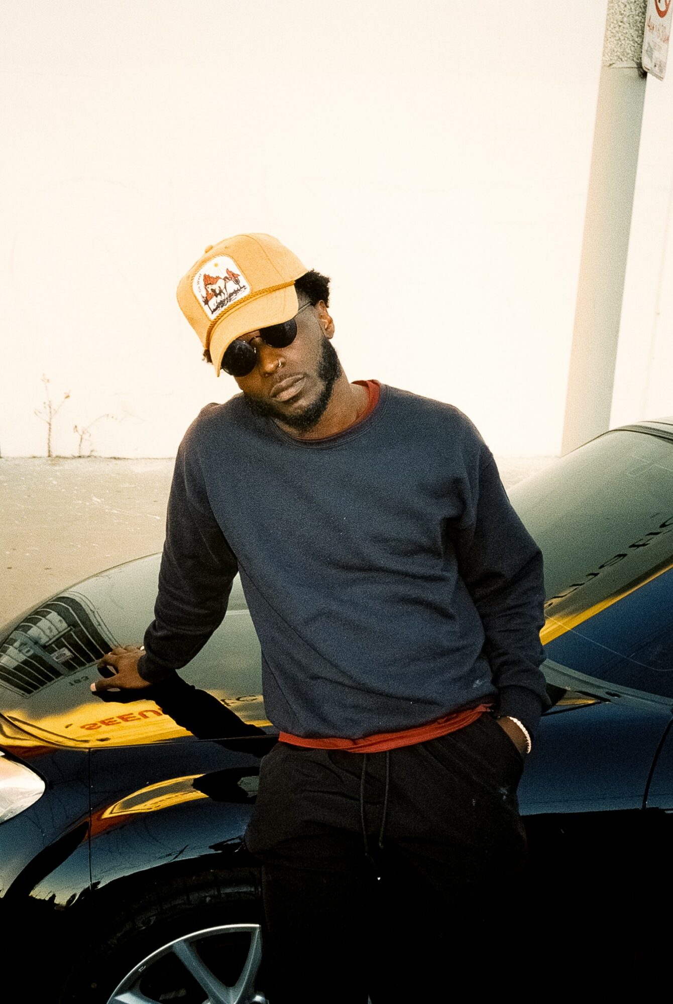 Person wearing sunglasses, a hat, and a dark sweater leaning on a black car against a plain background.
