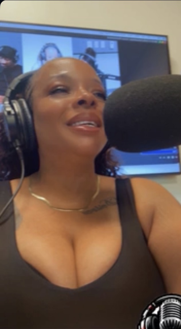 Woman speaking into a microphone with headphones, in a recording studio or radio station, smiling and looking slightly upward.