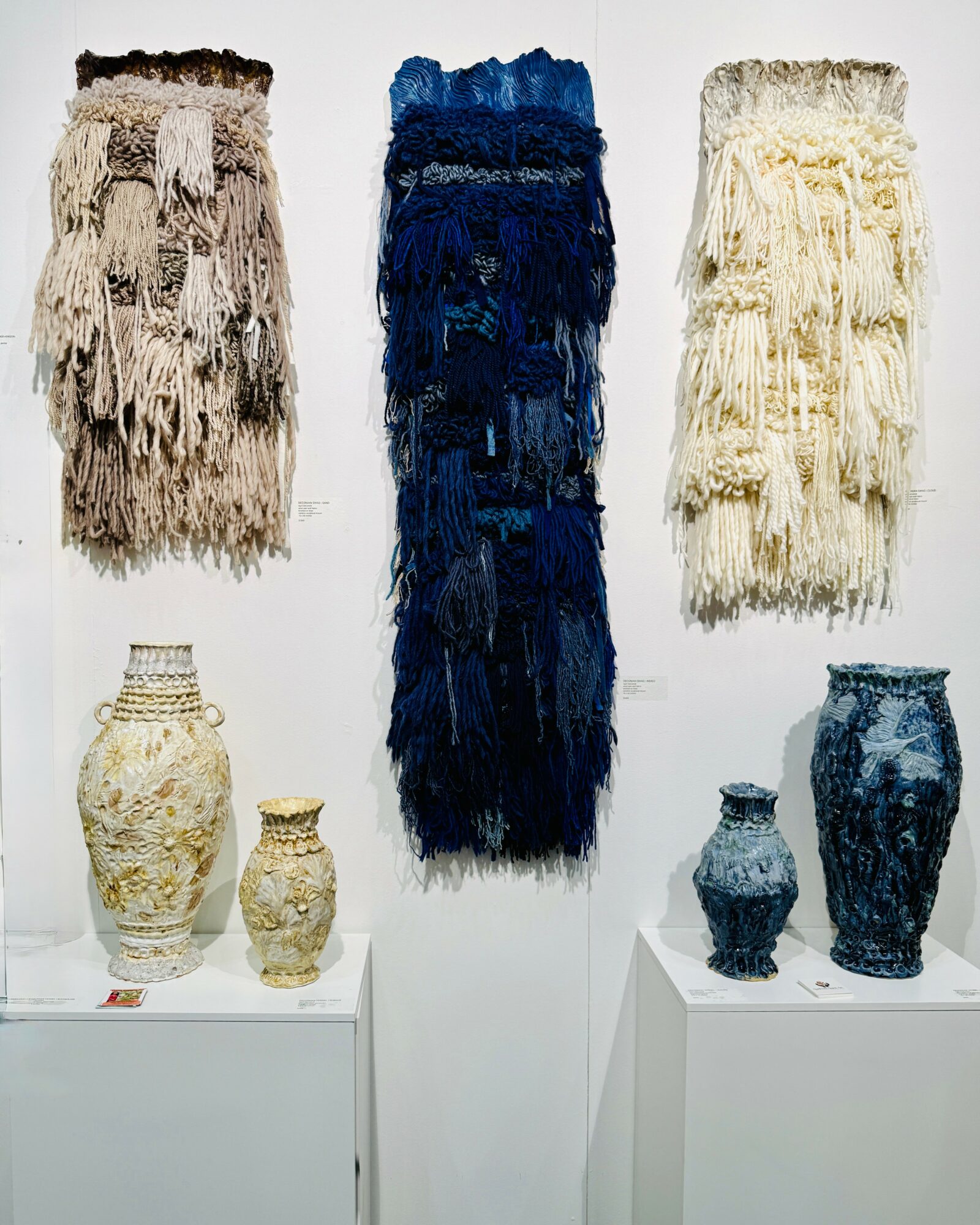 Three large textured wall hangings with fringe, and six vases of various sizes and colors displayed on white pedestals.
