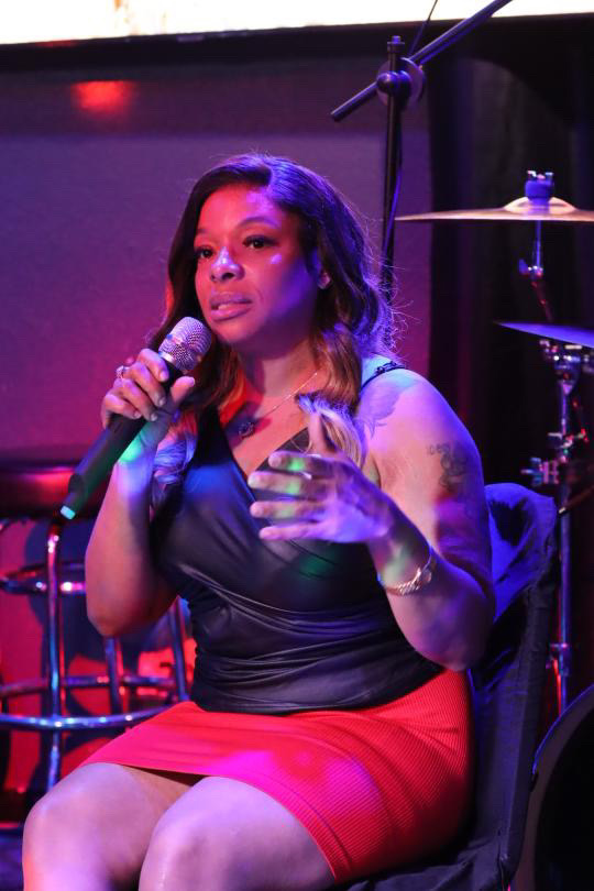 Woman singing into a microphone on stage, holding a glass, with musical instruments in background.